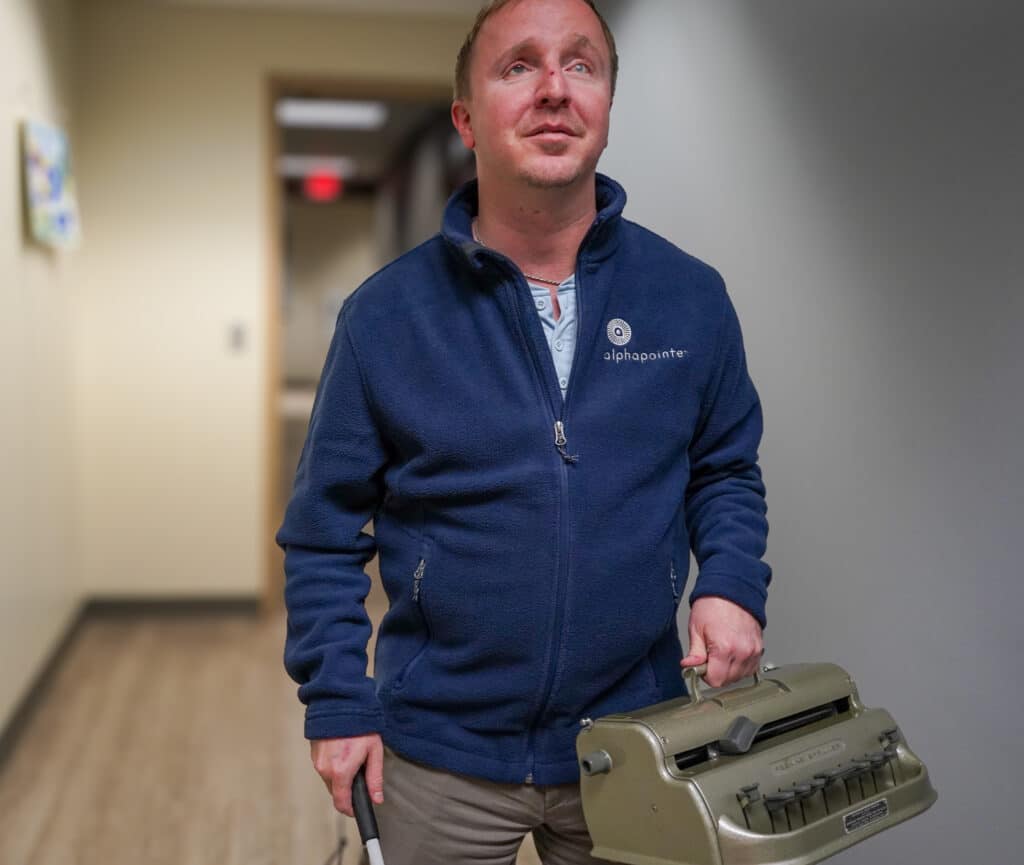 Cameron is carrying a Braille writer down the hallways of Alphapointe. 