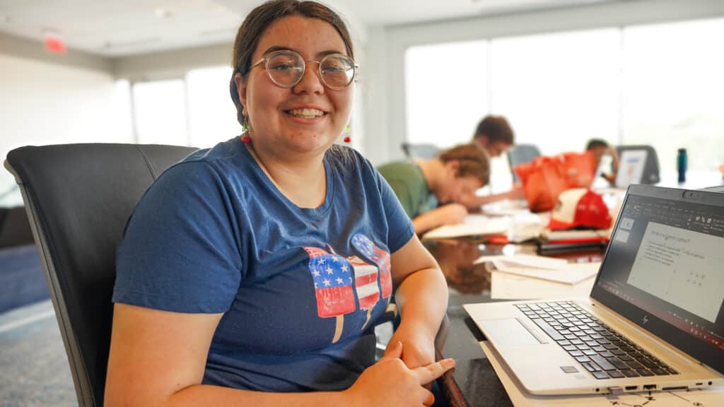 Liberty is facing the camera and smiling. She is seated in front of a computer at Technology Camp.