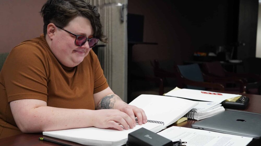 RJ is using both hands to run across a book of Braille