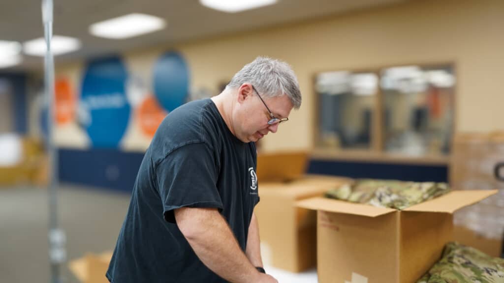 Marco is standing at a table folding pants and placing them in a box. 