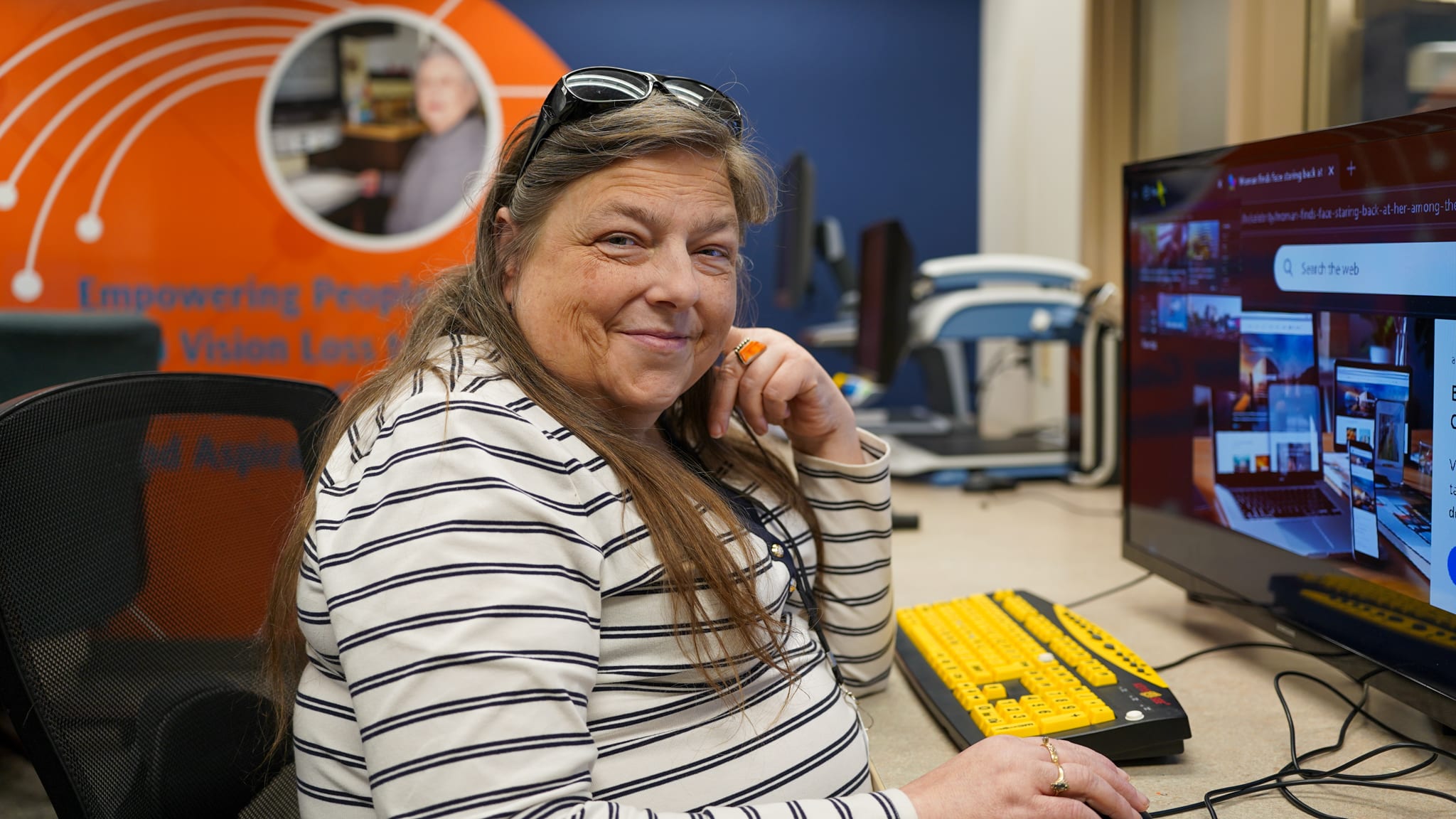 Emily is seated at a computer and is smiling at the camera. She is in our adaptive technology lab learning how to use different tools to use a computer.