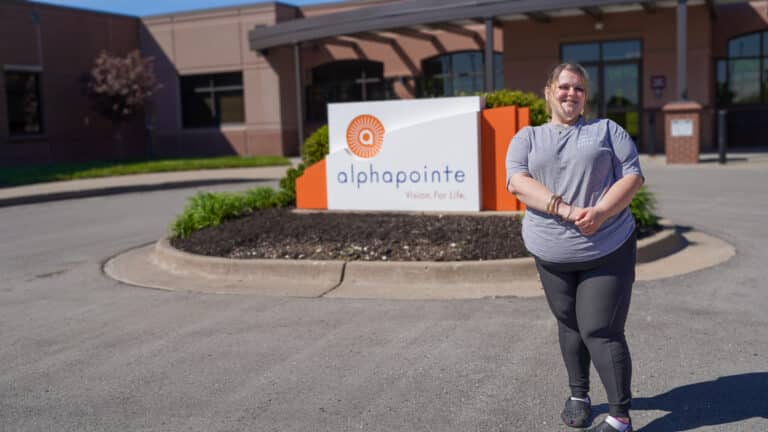 Olivia is standing out front of the Alphapointe building next to the white and orange Alphapointe sign. She is smiling at the camera.