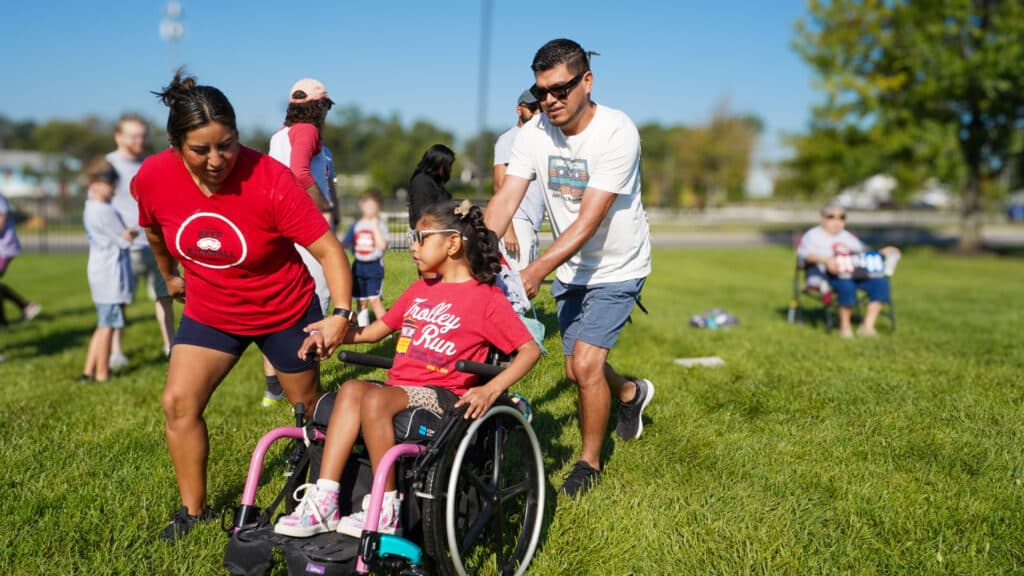 Volunteers help push a Beep Kickball participant who is in a wheelchair toward base.