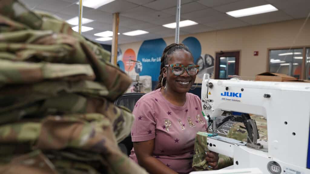 Tiffany is seated at a sewing machine surrounded by stacks of camouflage pants. She is smiling at the camera.