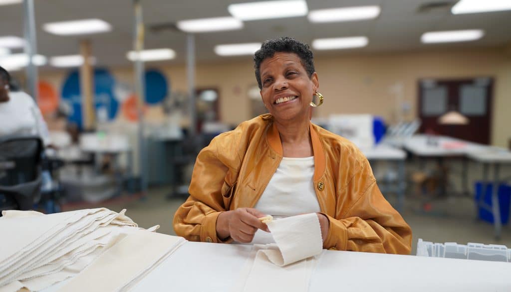 Robin is seated at a table where she works on sewing projects. She is smiling at the camera.