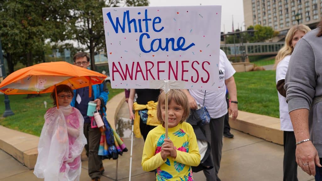 A young girl is walking with a group through the city. She is holding a sign that says "white cane awareness."