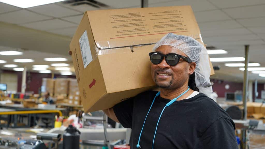 Romia (Ro-MEE) is carrying a box of caps after the have been printed on. He is wearing a hair net and dark glasses while walking through our Office Products department with a smile on his face.